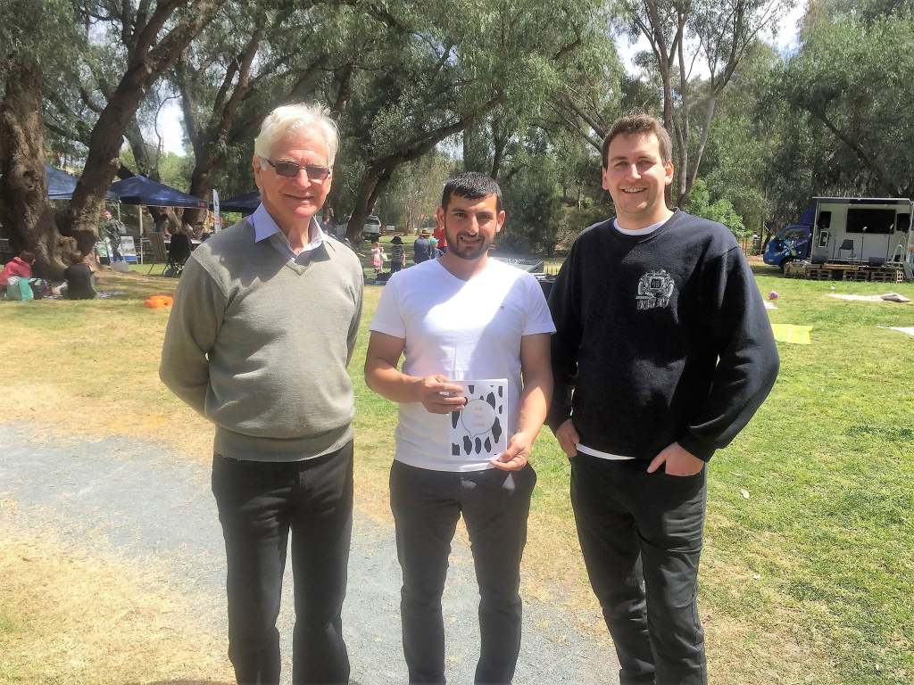 Hussam Saraf holding Ask the Cows beside two people at an outdoor community gathering.