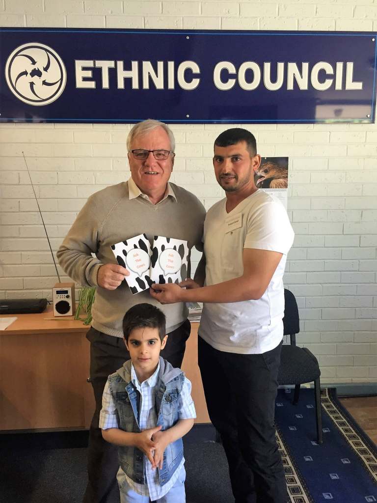 Hussam Saraf with an adult and child, holding Ask the Cows beneath an Ethnic Council sign.