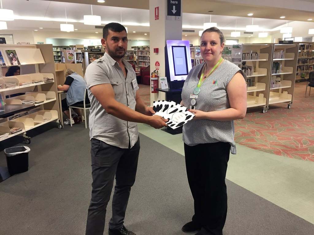 Hussam Saraf and a library staff member holding copies of Ask the Cows beside library shelves.