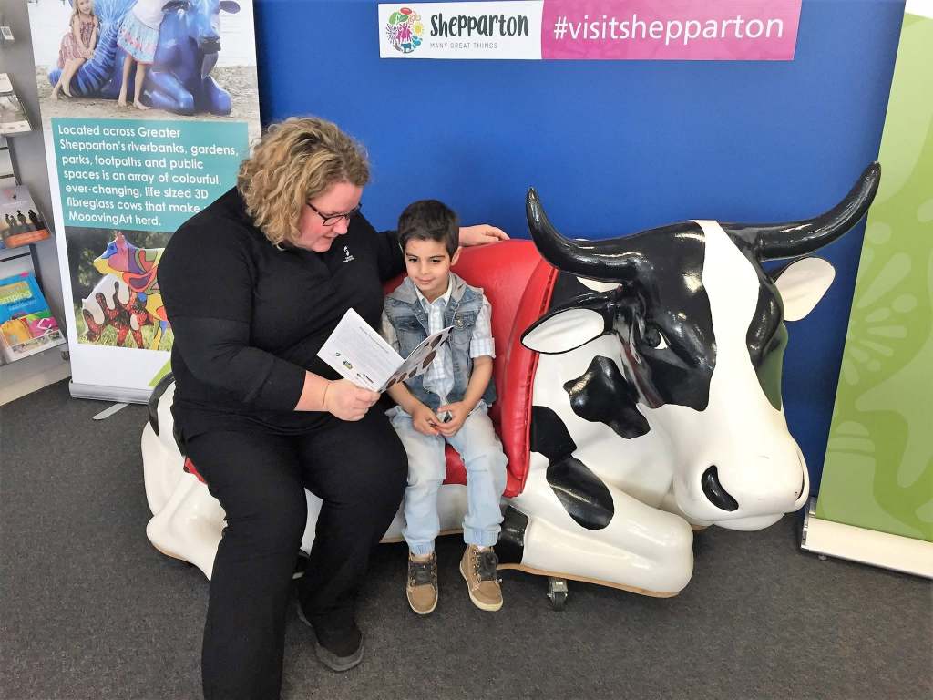 An adult and child reading together beside a cow sculpture and a Visit Shepparton display.