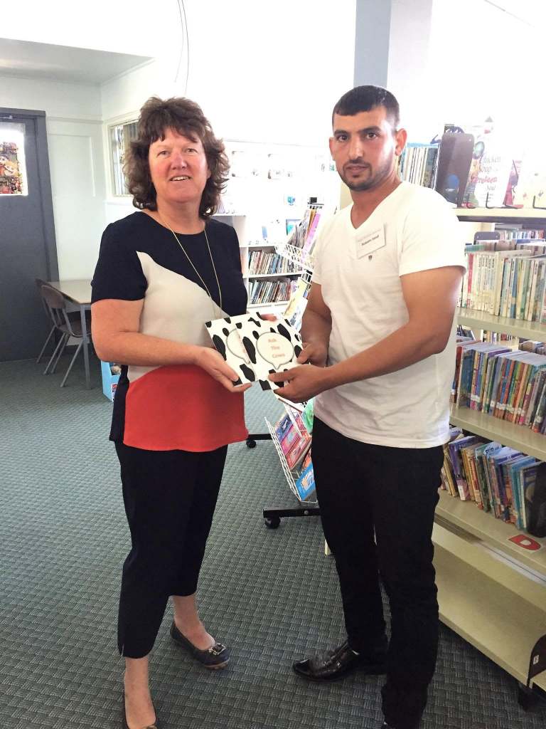 Hussam Saraf and a library staff member holding copies of Ask the Cows among bookshelves.