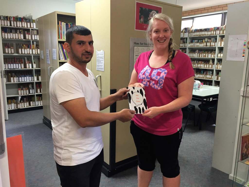 Hussam Saraf and a staff member holding Ask the Cows in front of library shelving.
