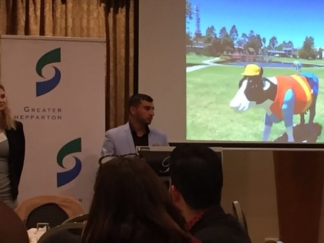 Hussam Saraf presenting beside a Greater Shepparton banner and a projected photograph of a colourful cow sculpture.