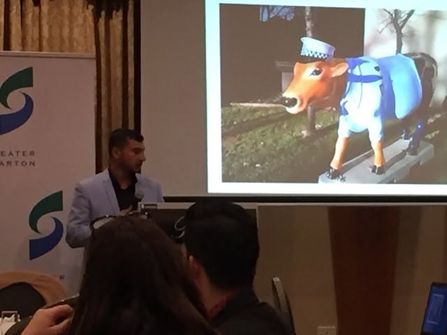 Hussam Saraf at a lectern beside a projected photograph of a cow sculpture dressed as a police officer.