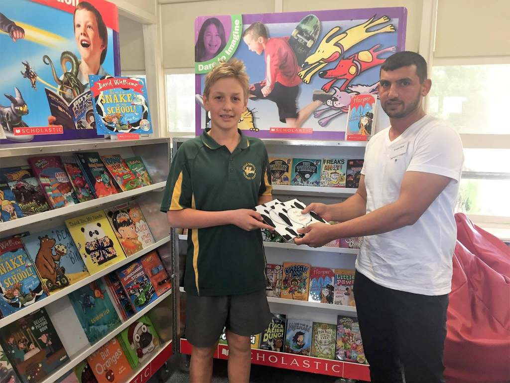 Hussam Saraf and a student in a green school uniform holding Ask the Cows beside children's books.