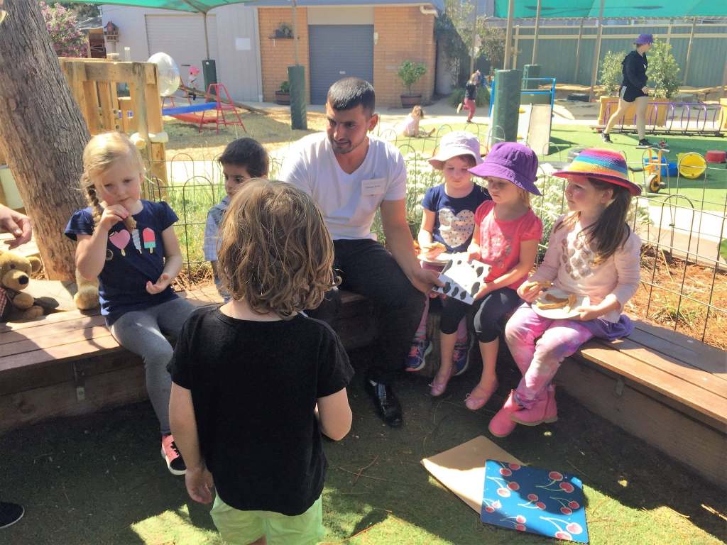 Hussam Saraf joining an adult and a group of children for story time in an outdoor play area.