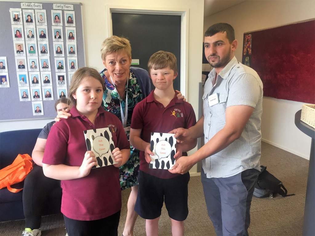 Hussam Saraf with an adult and two students in maroon school uniforms holding Ask the Cows.
