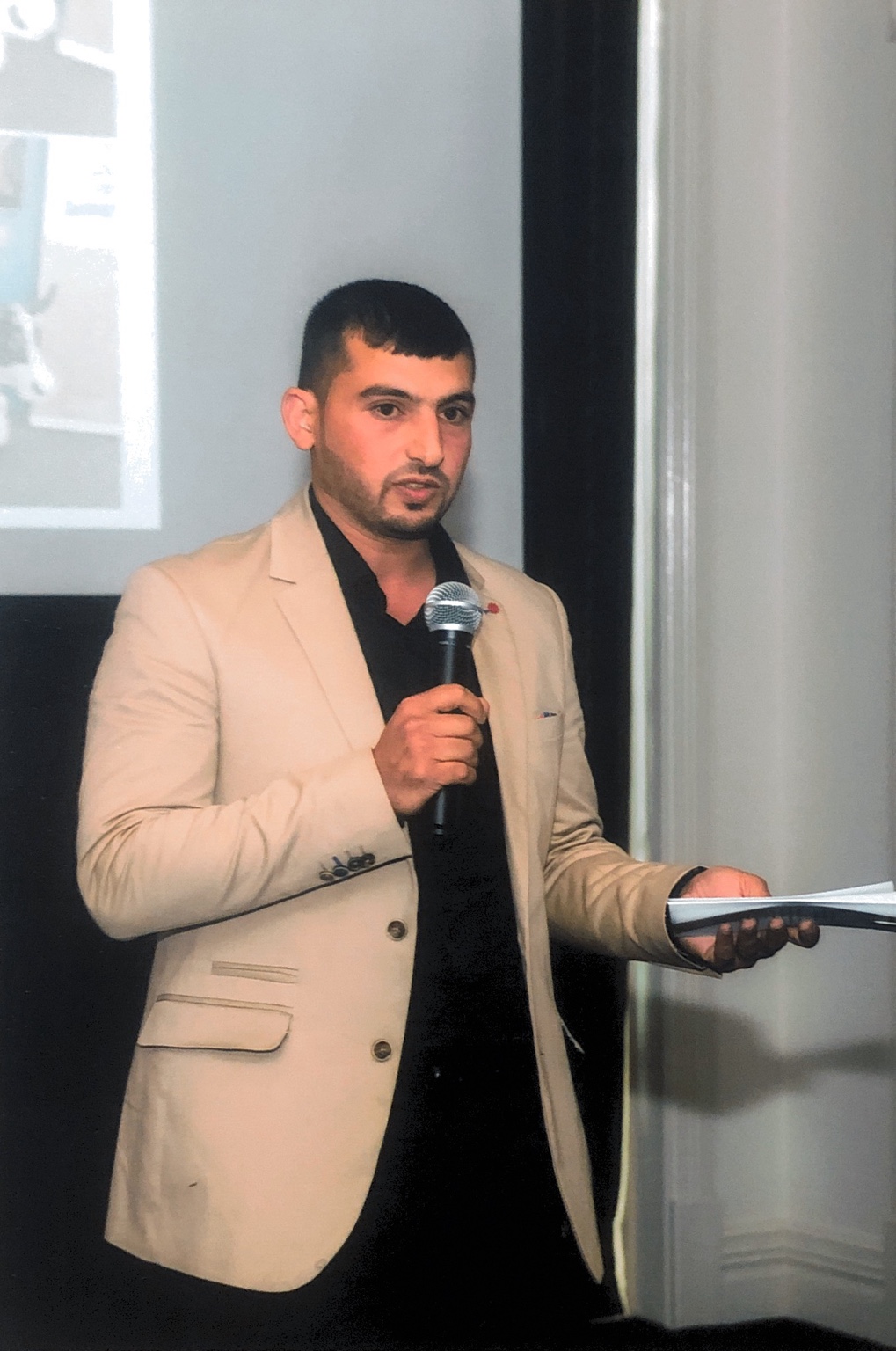 Hussam Saraf holding a microphone and notes while addressing the 2017 graduation gathering.