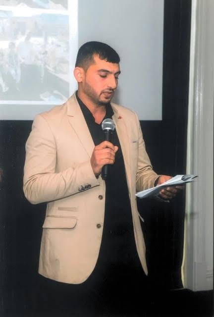 Hussam Saraf holding a microphone and notes while addressing the graduation gathering.