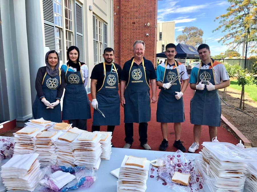 Six school and community volunteers wearing Rotary Mooroopna aprons behind a food table.