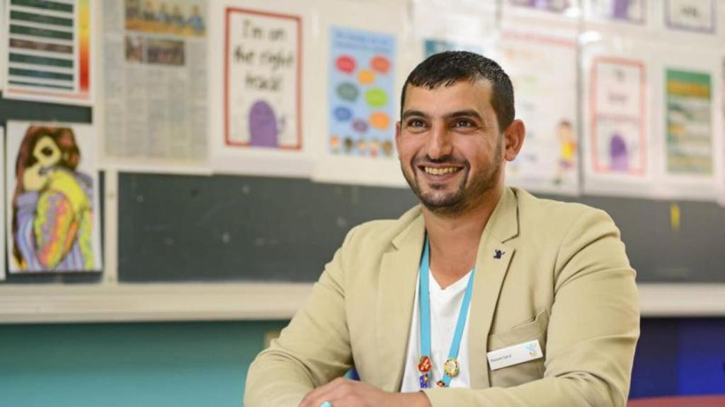 Hussam Saraf smiling in a classroom, wearing a beige jacket and a staff lanyard.