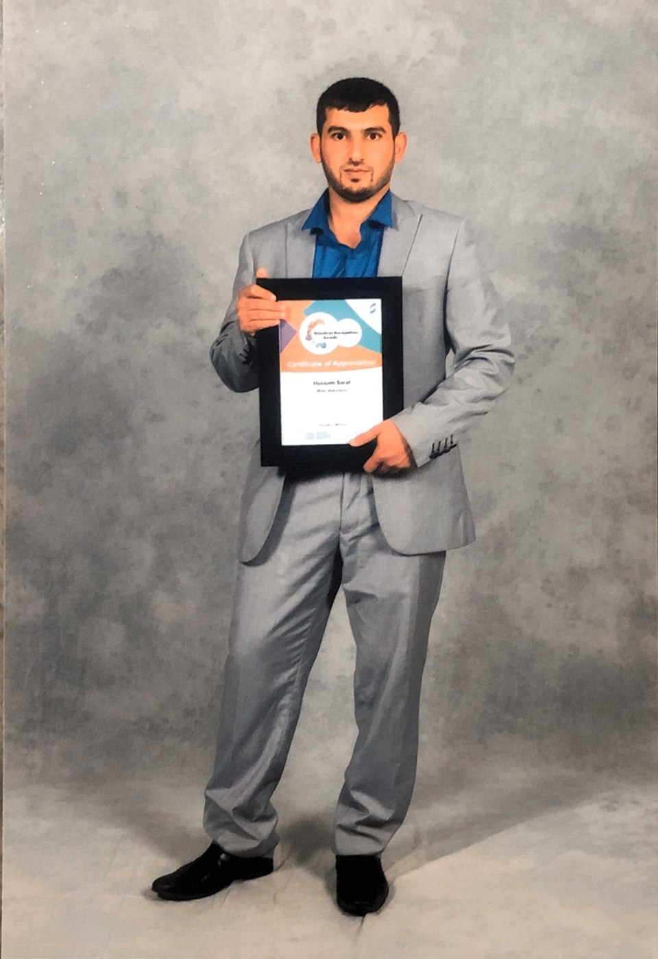 A full-length portrait of Hussam Saraf holding his Male Volunteer Certificate of Appreciation in 2017.