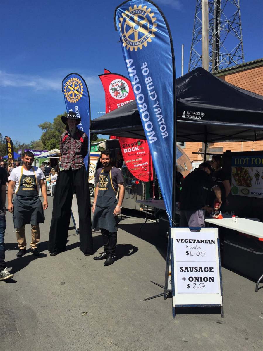Rotary volunteers beside club flags and a stilt performer at the food festival.