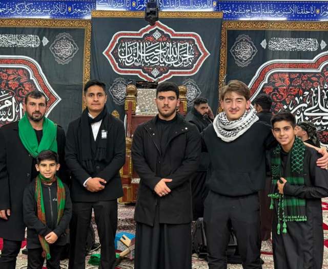 A group of community members gathered indoors in front of Ashura commemorative banners.